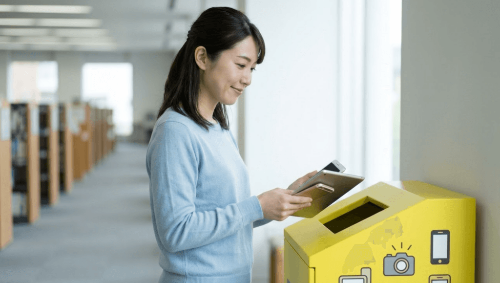 自治体の施設に設置された小型家電回収ボックスに、古いタブレットを投入しようとする日本人女性。