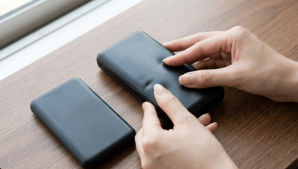 日本人女性の手が、木目のデスクの上に置かれた、わずかにケースが膨張している黒いモバイルバッテリーの側面を慎重に指で押して確認している様子。