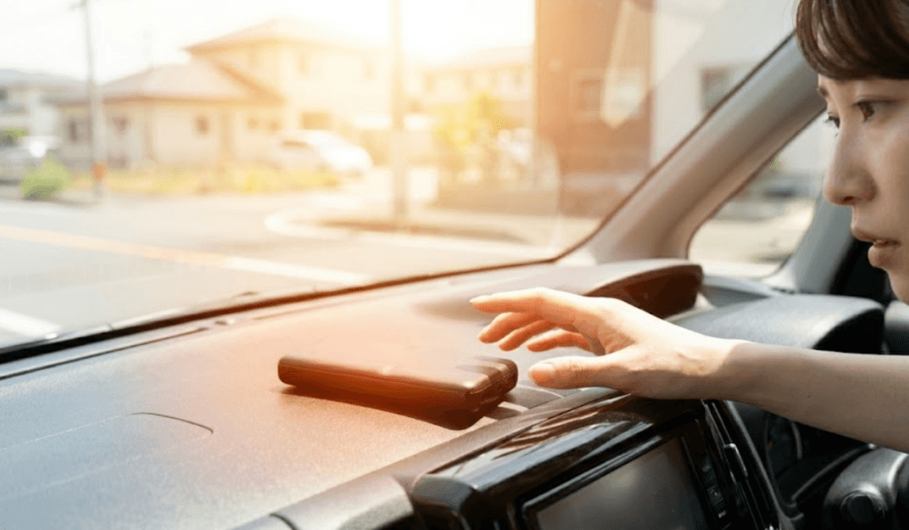 真夏の晴れた日に、強い日差しが差し込む車のダッシュボードの上に放置された黒いモバイルバッテリー。日本人女性の手がその熱さに気づき、注意深く手を伸ばしている。