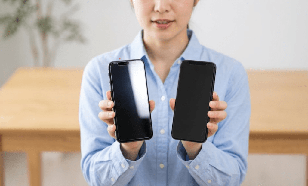 スマートフォンのガラスフィルムとPETフィルムの厚みの違いを比較する女性。左のスマートフォンは厚みのあるガラスフィルムが光を反射し、右は極薄のPETフィルムが装着されている。