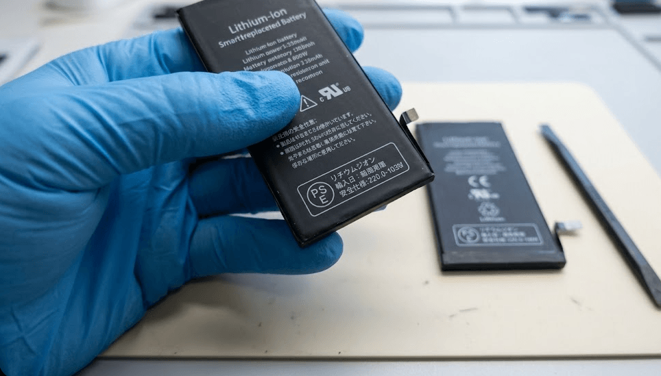 静電防止グローブを着用したアジア人作業員の手が、スマートフォン用交換バッテリーを持ち、ラベルに印字された丸形の「PSEマーク」と事業者名を指し示しているクローズアップ写真。デスク上には修理用具が並ぶ。